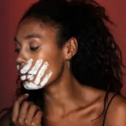 Woman with white cream on her face, applying it to her face against a maroon background